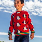 Child wearing the Frugi scarlet penguins organic cotton jolly knitted jumper. Red knitted jumper with repeat penguin print and rainbow stripe hemline and arm cuffs with denim jeans.