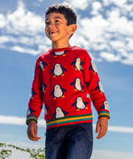 Child wearing the Frugi scarlet penguins organic cotton jolly knitted jumper. Red knitted jumper with repeat penguin print and rainbow stripe hemline and arm cuffs with denim jeans.