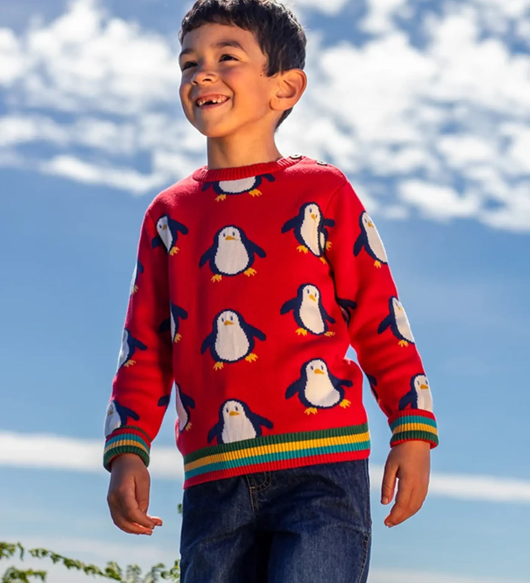 Child wearing the Frugi scarlet penguins organic cotton jolly knitted jumper. Red knitted jumper with repeat penguin print and rainbow stripe hemline and arm cuffs with denim jeans.