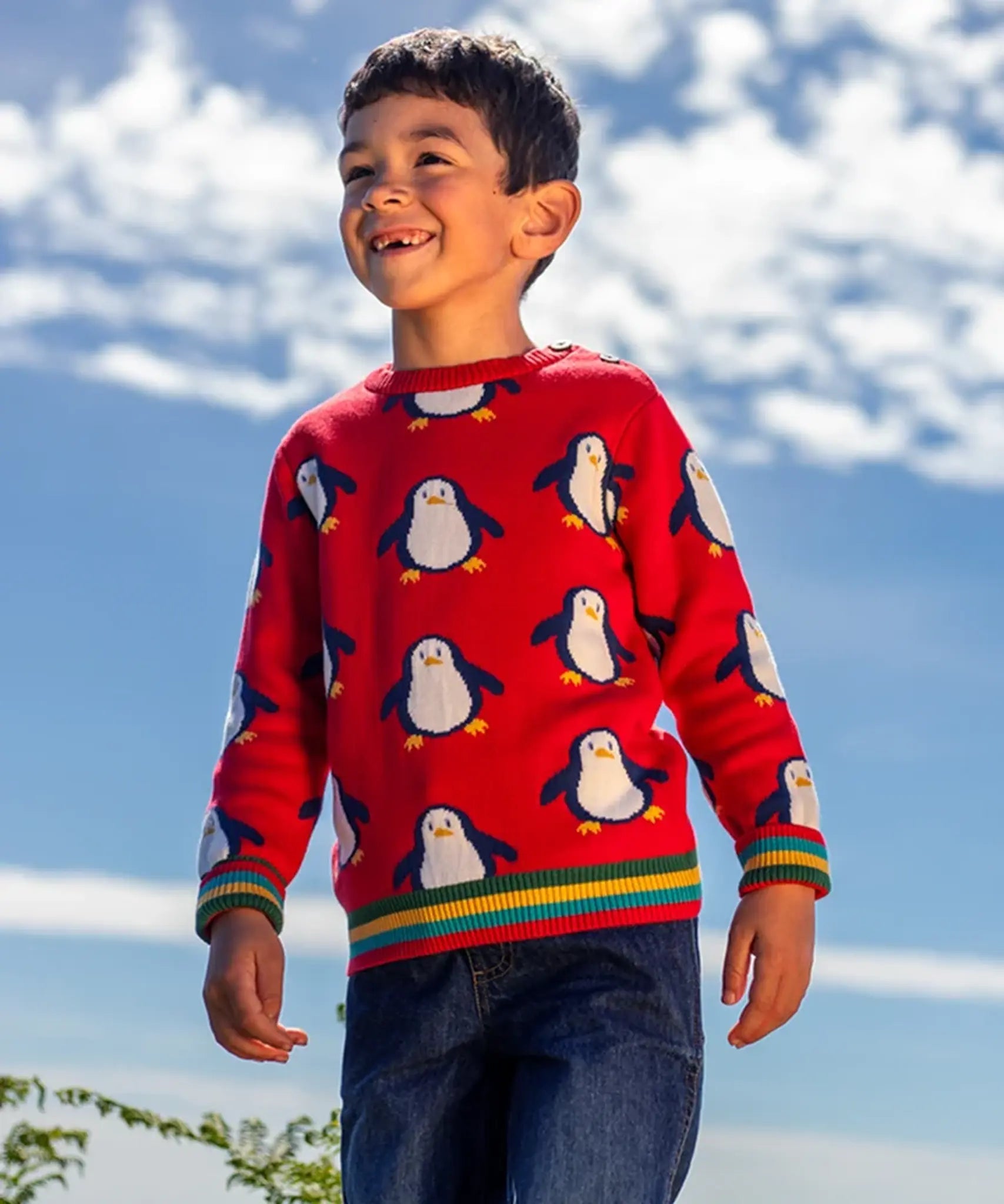 Child wearing the Frugi scarlet penguins organic cotton jolly knitted jumper. Red knitted jumper with repeat penguin print and rainbow stripe hemline and arm cuffs with denim jeans.