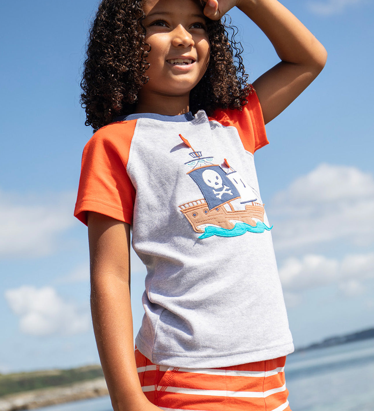 A smiling young child wearing the Frugi Renny Raglan T-Shirt in Grey Marl, featuring an embroidered pirate ship design in the centre, and orange sleeves with a navy collar, stood on a pebbled beach.