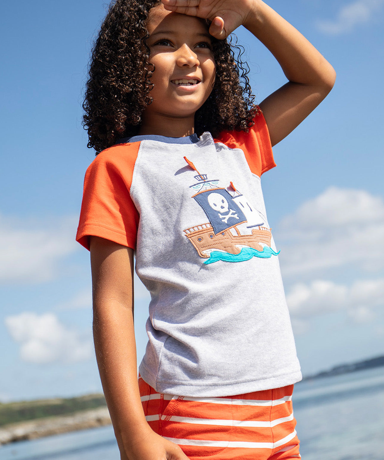 A smiling young child wearing the Frugi Renny Raglan T-Shirt in Grey Marl, featuring an embroidered pirate ship design in the centre, and orange sleeves with a navy collar, stood on a pebbled beach.