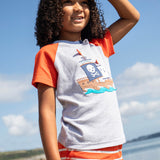 A smiling young child wearing the Frugi Renny Raglan T-Shirt in Grey Marl, featuring an embroidered pirate ship design in the centre, and orange sleeves with a navy collar, stood on a pebbled beach.