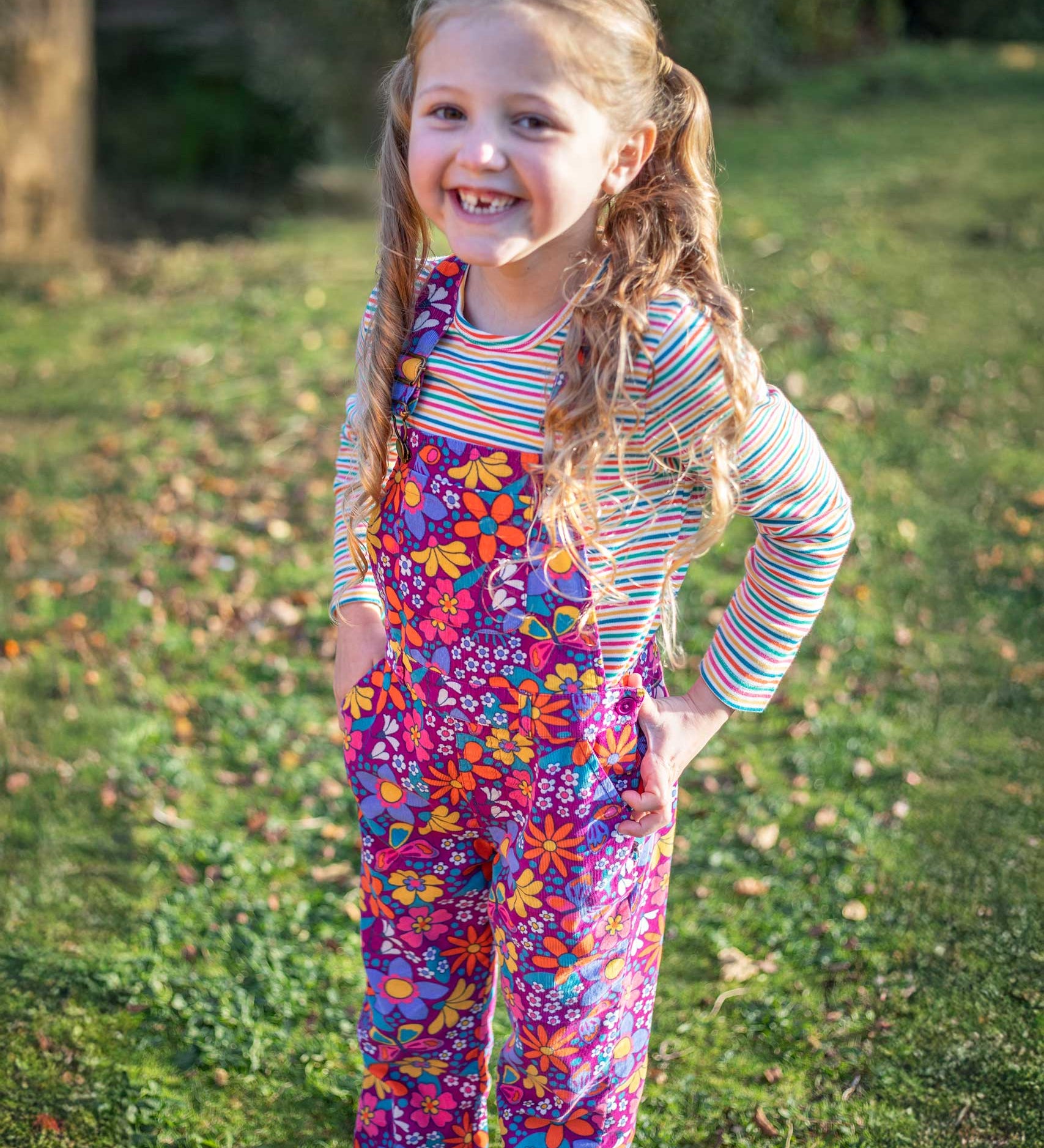 A child wearing the Frugi Retro Floral Kids' Flared Organic cotton Corduroy Dungarees with a long sleeved rainbow stripe top underneath. These cord dungarees are part of a wide range of organic cotton clothing for children available here at Babipur.