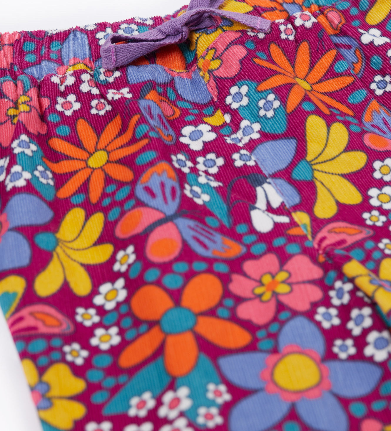 A close up of the purple waist tie on the Frugi Retro Floral print Nieva Cotton Corduroy Trousers. A purple coloured corduroy with a colourful all over floral print. These trousers are part of a wide range of organic cotton clothing for children available here at Babipur.
