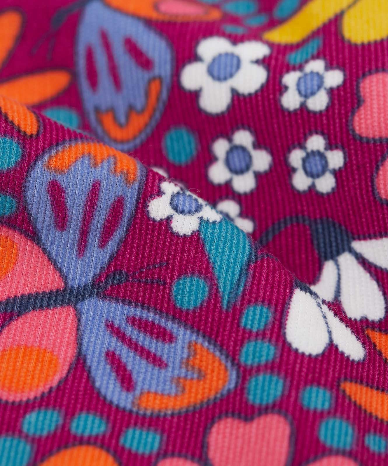 A close up view of the material of the Frugi Retro Floral print Nieva Cotton Corduroy Trousers. A purple coloured corduroy with a colourful all over floral print. These trousers are part of a wide range of organic cotton clothing for children available here at Babipur.