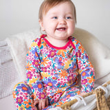 A baby sitting up wearing the Frugi easy dressing printed babygrow in white with aretro floral design. A GOTS organic cotton all in one with a bright retro floral design and pink trim around the neck and shoulder. This babygrow is a part of a wide range of organic cotton clothing for children available here at Babipur. 