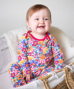 A baby sitting up wearing the Frugi easy dressing printed babygrow in white with aretro floral design. A GOTS organic cotton all in one with a bright retro floral design and pink trim around the neck and shoulder. This babygrow is a part of a wide range of organic cotton clothing for children available here at Babipur. 