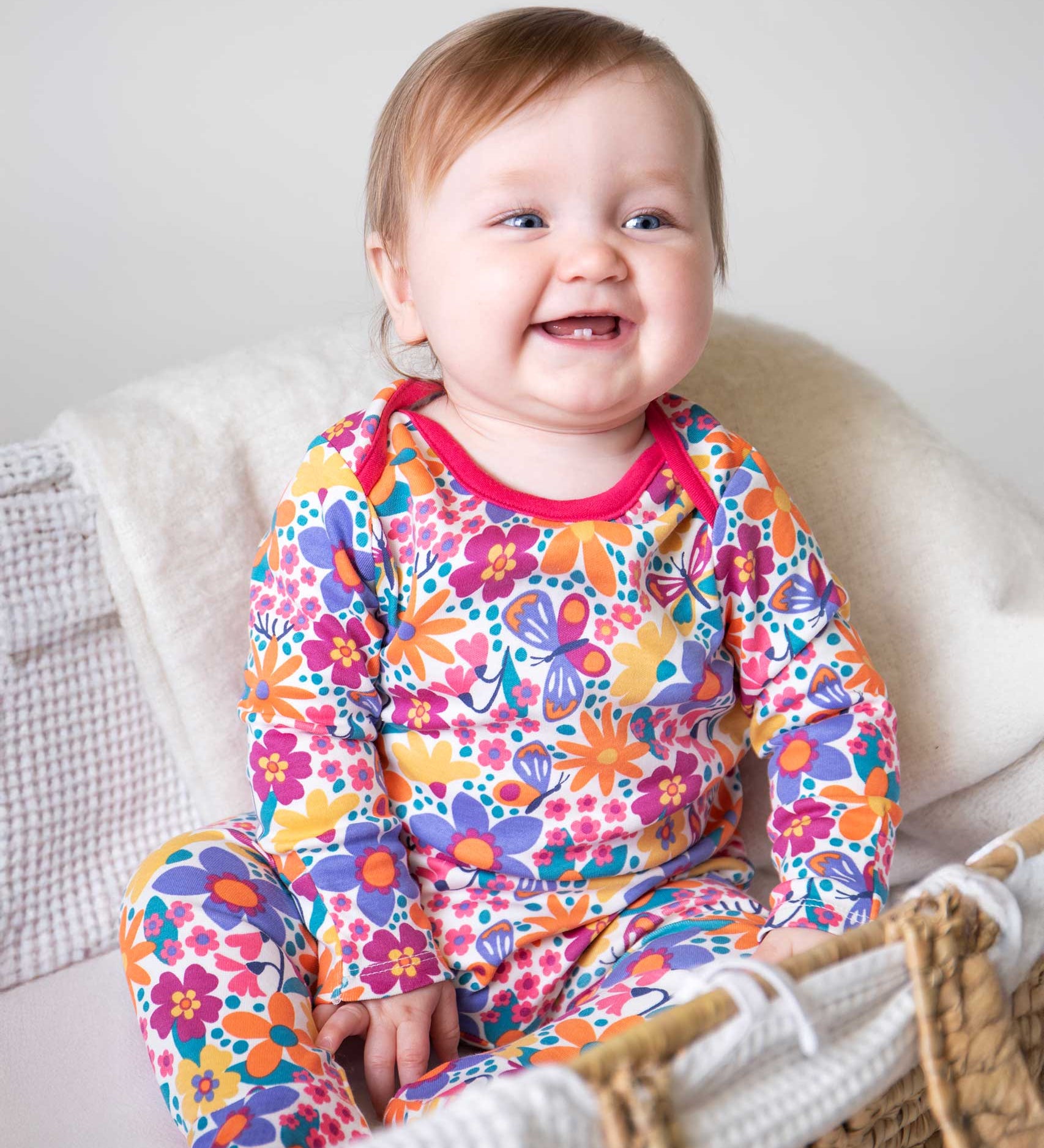 A baby sitting up wearing the Frugi easy dressing printed babygrow in white with aretro floral design. A GOTS organic cotton all in one with a bright retro floral design and pink trim around the neck and shoulder. This babygrow is a part of a wide range of organic cotton clothing for children available here at Babipur. 
