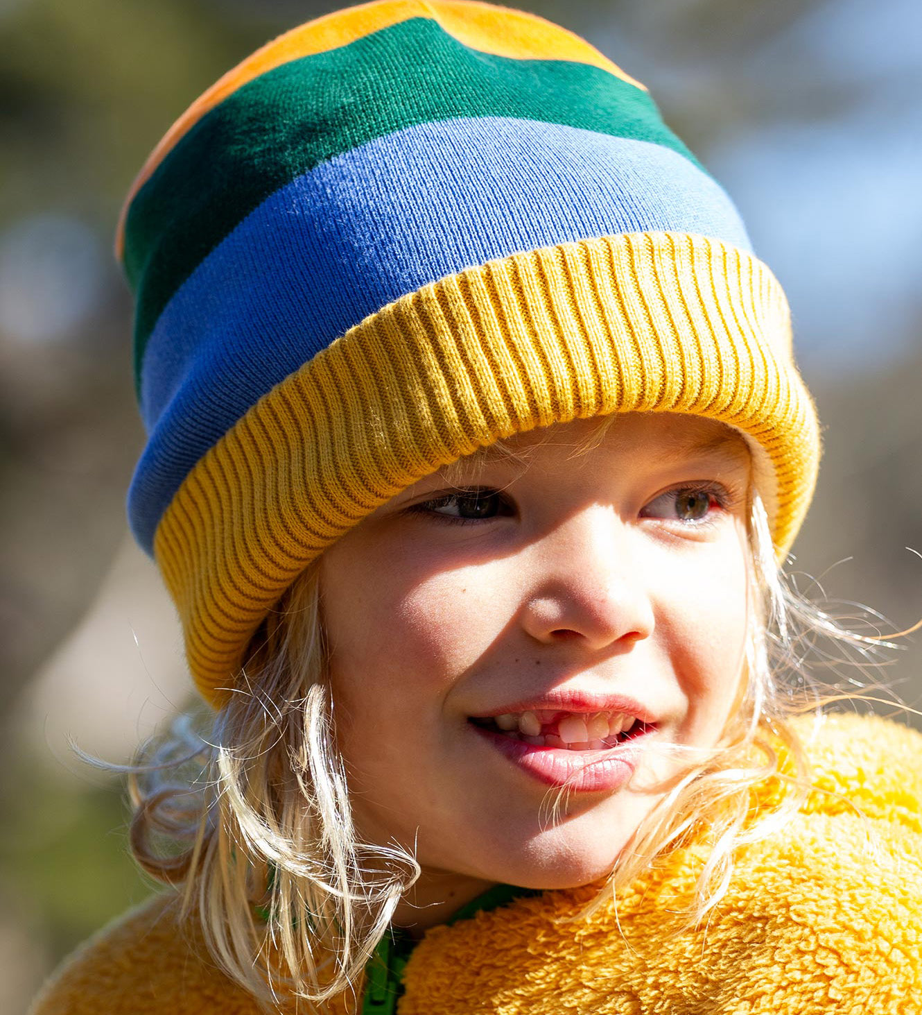 A child wearing the Frugi reversible voyager hat in penguin rainbow showing the bold yellow green and blue stripe design with a yellow soft ribbed trim.
