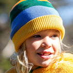 A child wearing the Frugi reversible voyager hat in penguin rainbow showing the bold yellow green and blue stripe design with a yellow soft ribbed trim.