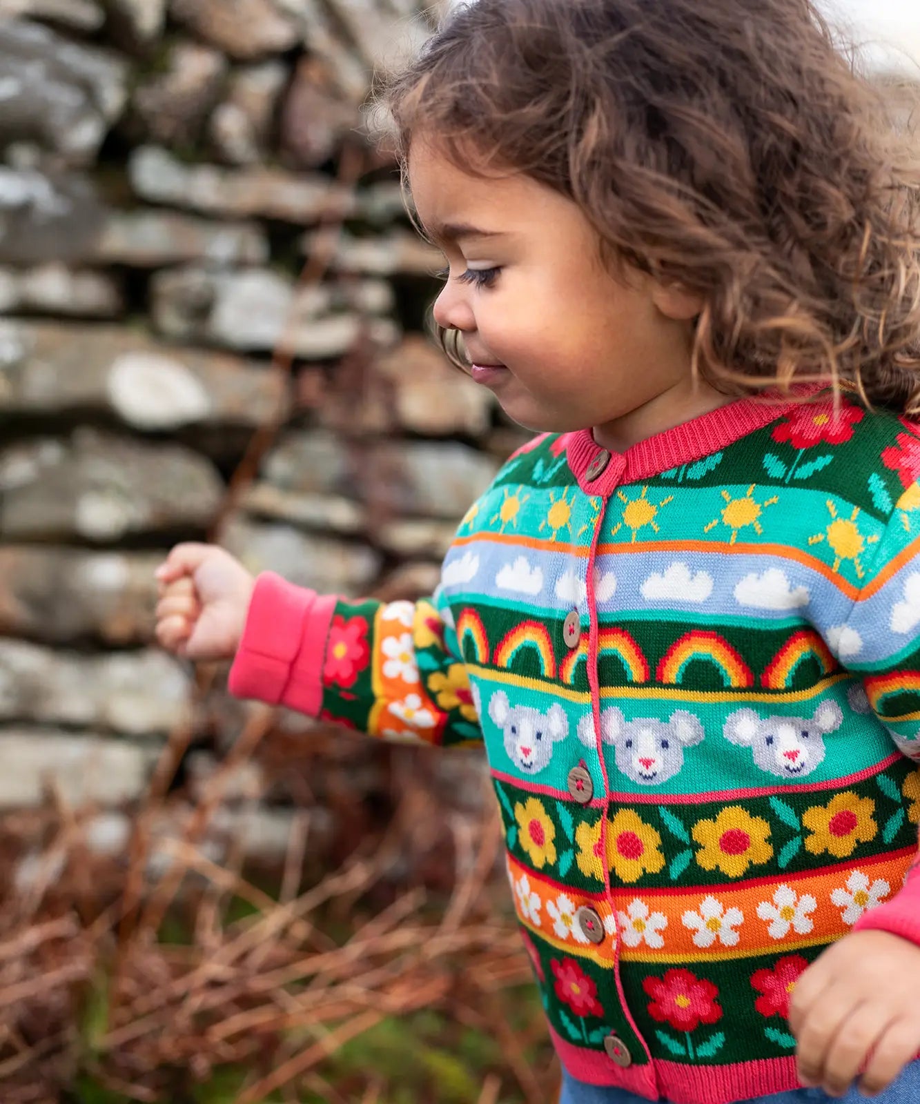Child wearing the Frugi Robyn fairisle cardigan with a mouse Fair Isle design featuring flowers, rainbows, clouds and suns. 
