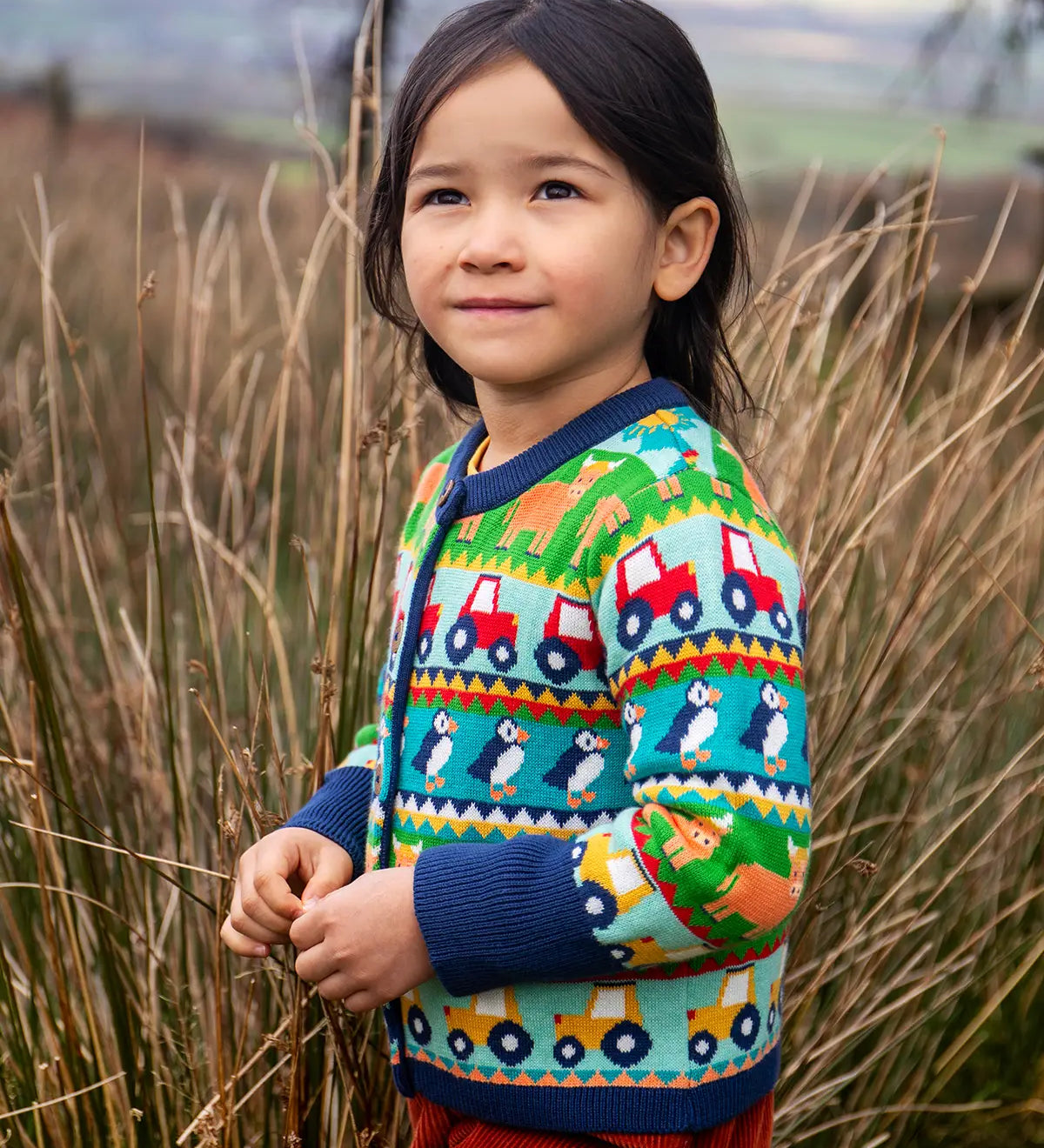Child wearing the Frugi Robyn fairisle cardigan with colourful farm design featuring tractors, highland coos and puffins. Button up front and navy cuffs, neck and waist band. 