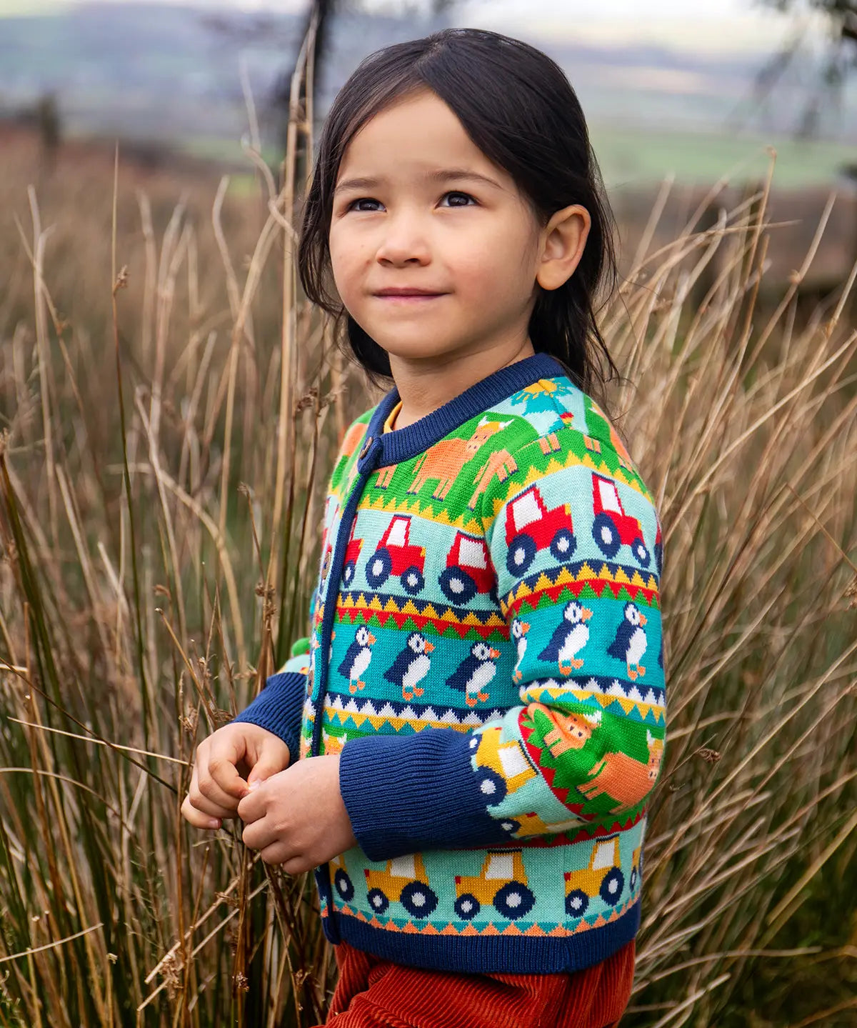 Child wearing the Frugi Robyn fairisle cardigan with colourful farm design featuring tractors, highland coos and puffins. Button up front and navy cuffs, neck and waist band. 