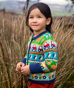 Child wearing the Frugi Robyn fairisle cardigan with colourful farm design featuring tractors, highland coos and puffins. Button up front and navy cuffs, neck and waist band. 