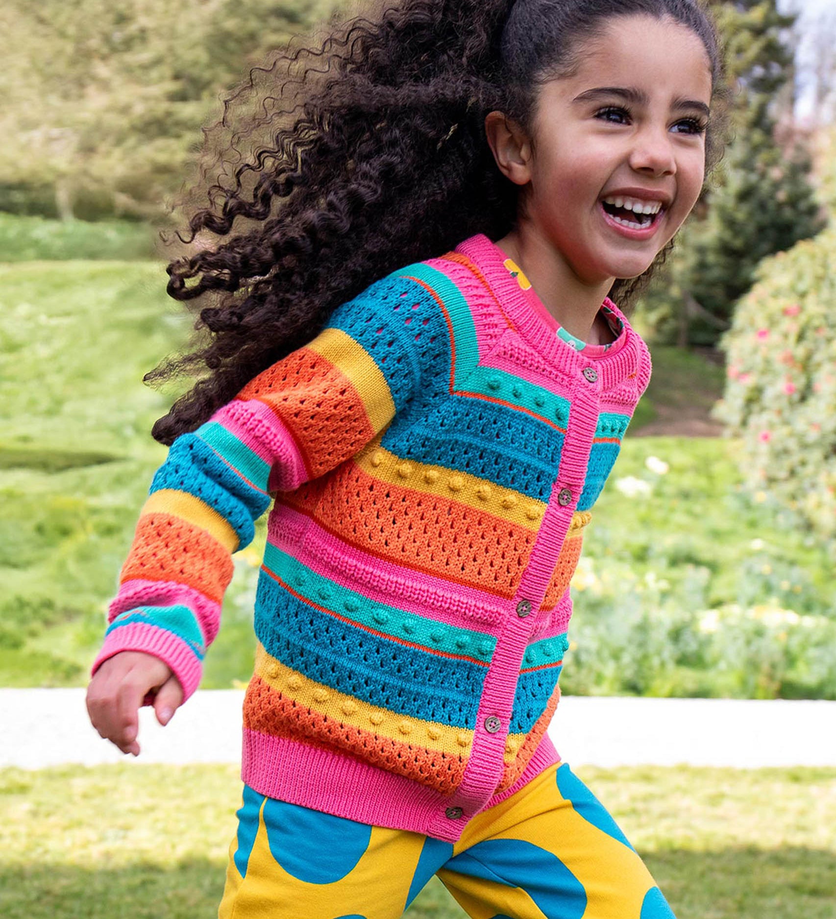 A child running in a park and wearing the Frugi kids rowe rainbow cardigan in rainbow/pointelle.