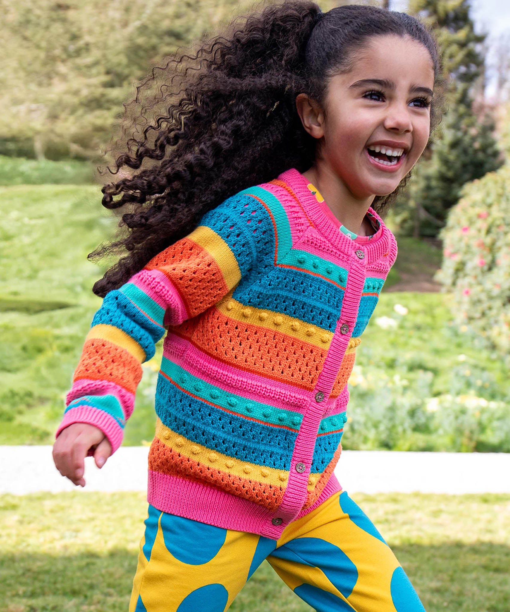 A child running in a park and wearing the Frugi kids rowe rainbow cardigan in rainbow/pointelle.
