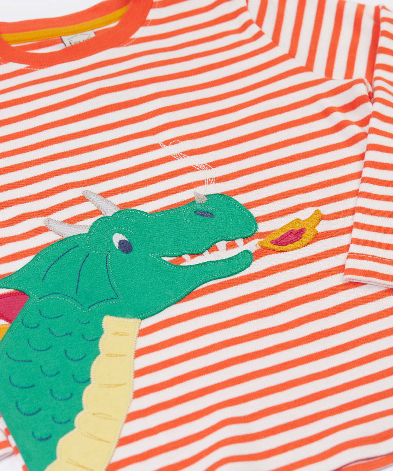 A close up of the fire breathing dragon on the front of the Frugi Salamander Stripe Dragon Discovery Circular Top. This orange and white striped top has a green coloured dragon applique detail on the front. This top is a part of a wide range of organic cotton clothing for children available here at Babipur. 
