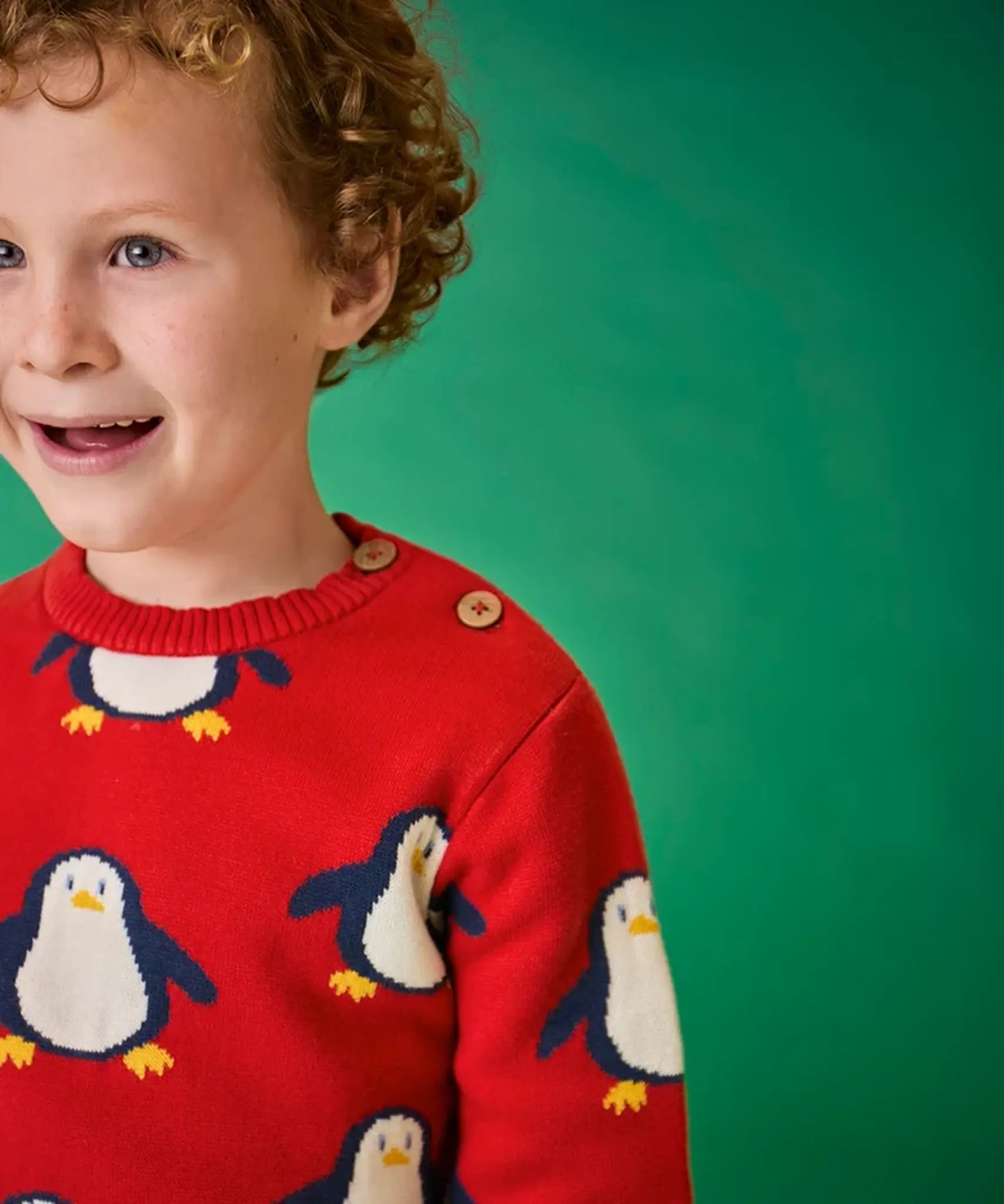Child wearing the Frugi scarlet penguins organic cotton jolly knitted jumper. Red knitted jumper with repeat penguin print and rainbow stripe hemline and arm cuffs. Close up of the button closure on the shoulder. 