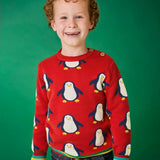 Child wearing the Frugi scarlet penguins organic cotton jolly knitted jumper. Red knitted jumper with repeat penguin print and rainbow stripe hemline and arm cuffs with denim jeans.
