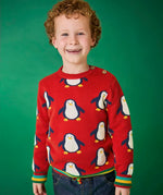 Child wearing the Frugi scarlet penguins organic cotton jolly knitted jumper. Red knitted jumper with repeat penguin print and rainbow stripe hemline and arm cuffs with denim jeans.