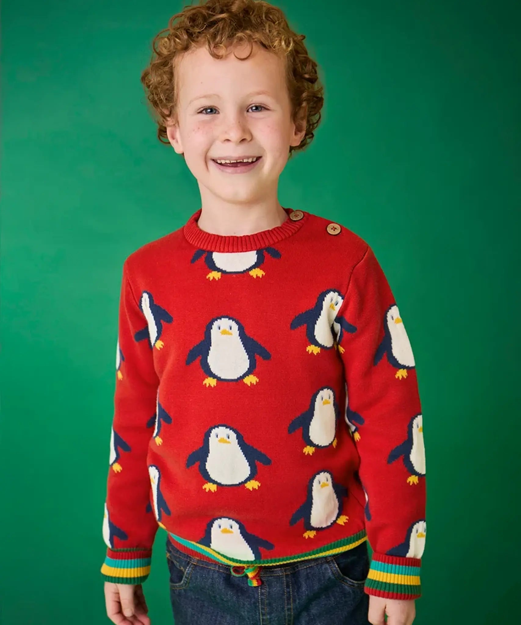 Child wearing the Frugi scarlet penguins organic cotton jolly knitted jumper. Red knitted jumper with repeat penguin print and rainbow stripe hemline and arm cuffs with denim jeans.