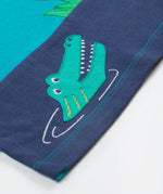 A close up of the Frugi sea, land, canopy short sleeve -shirt. Showing the crocodil applique and light blue stitching on the hem of the t-shirt on blue organic cotton fabric