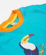 A close up of the Frugi sea, land, canopy short sleeve -shirt. Showing the shoulder fastener, orange neck trim and the toucan applique stitched on a blue organic cotton fabric