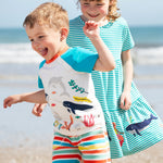 A child wearing the Frugi sea life Organic Reid Raglan T-Shirt with brightly coloured striped shorts. There is another child standing behind them wearing a blue and white striped dress.