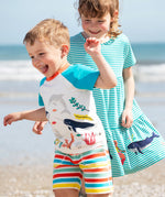 A child wearing the Frugi sea life Organic Reid Raglan T-Shirt with brightly coloured striped shorts. There is another child standing behind them wearing a blue and white striped dress.