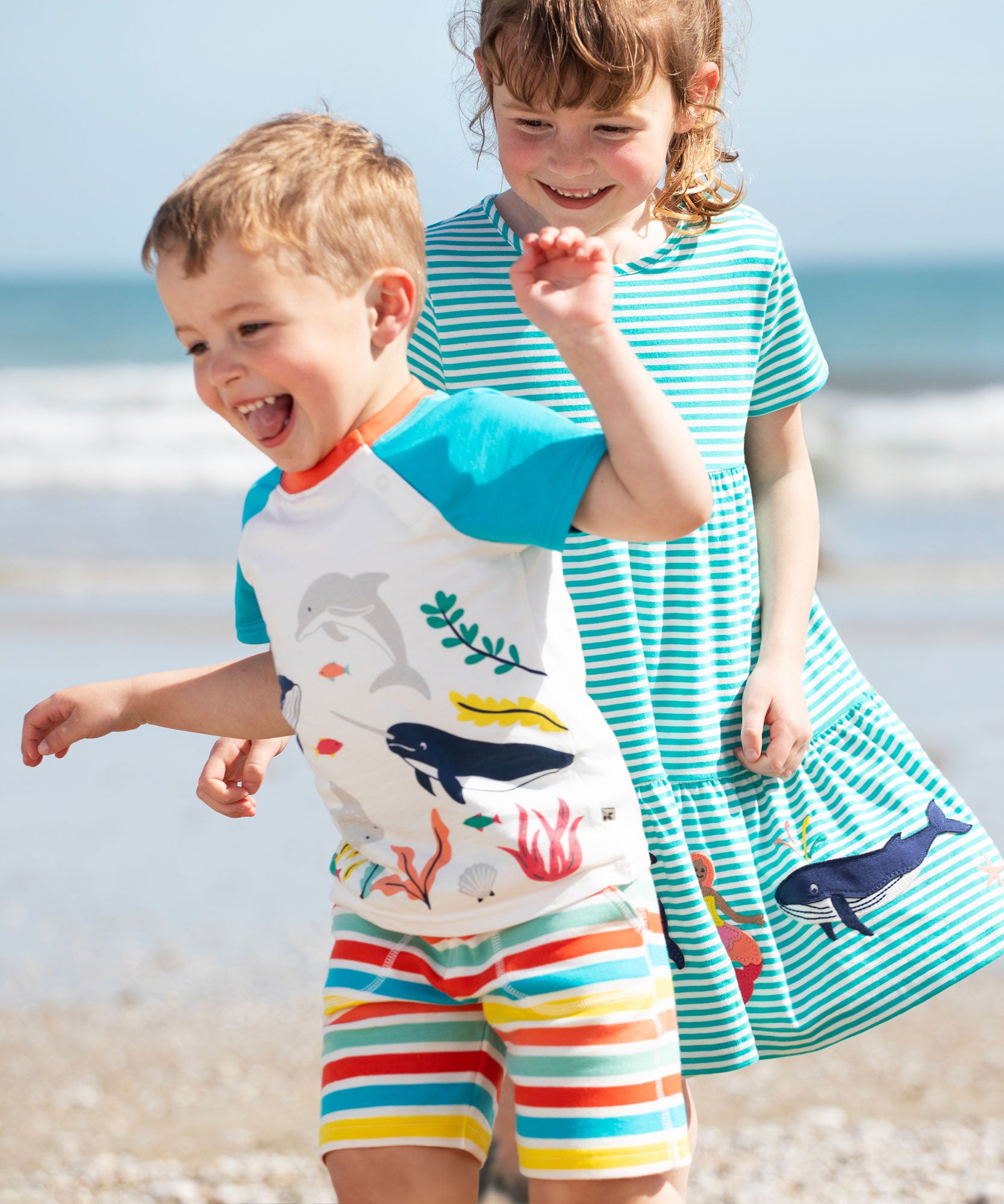 A child wearing the Frugi sea life Organic Reid Raglan T-Shirt with brightly coloured striped shorts. There is another child standing behind them wearing a blue and white striped dress.