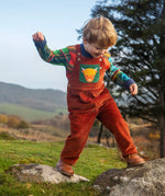 Child wearing the Frugi sebby brown rust organic cotton cord dungaree with a green chest pocket featuring a highland cow applique with a knitted jumper underneath.