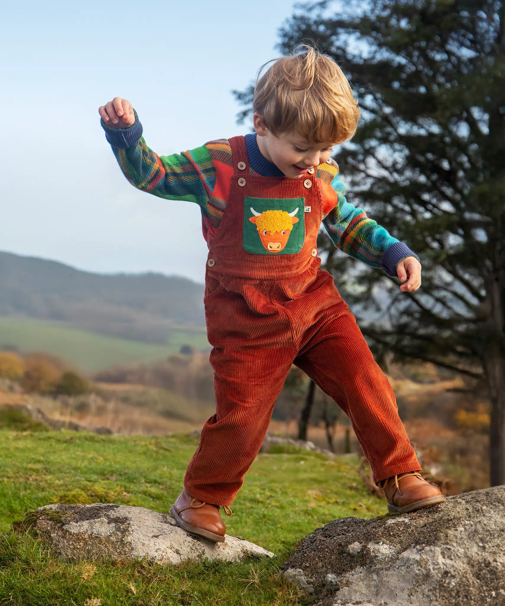 Child wearing the Frugi sebby brown rust organic cotton cord dungaree with a green chest pocket featuring a highland cow applique with a knitted jumper underneath.