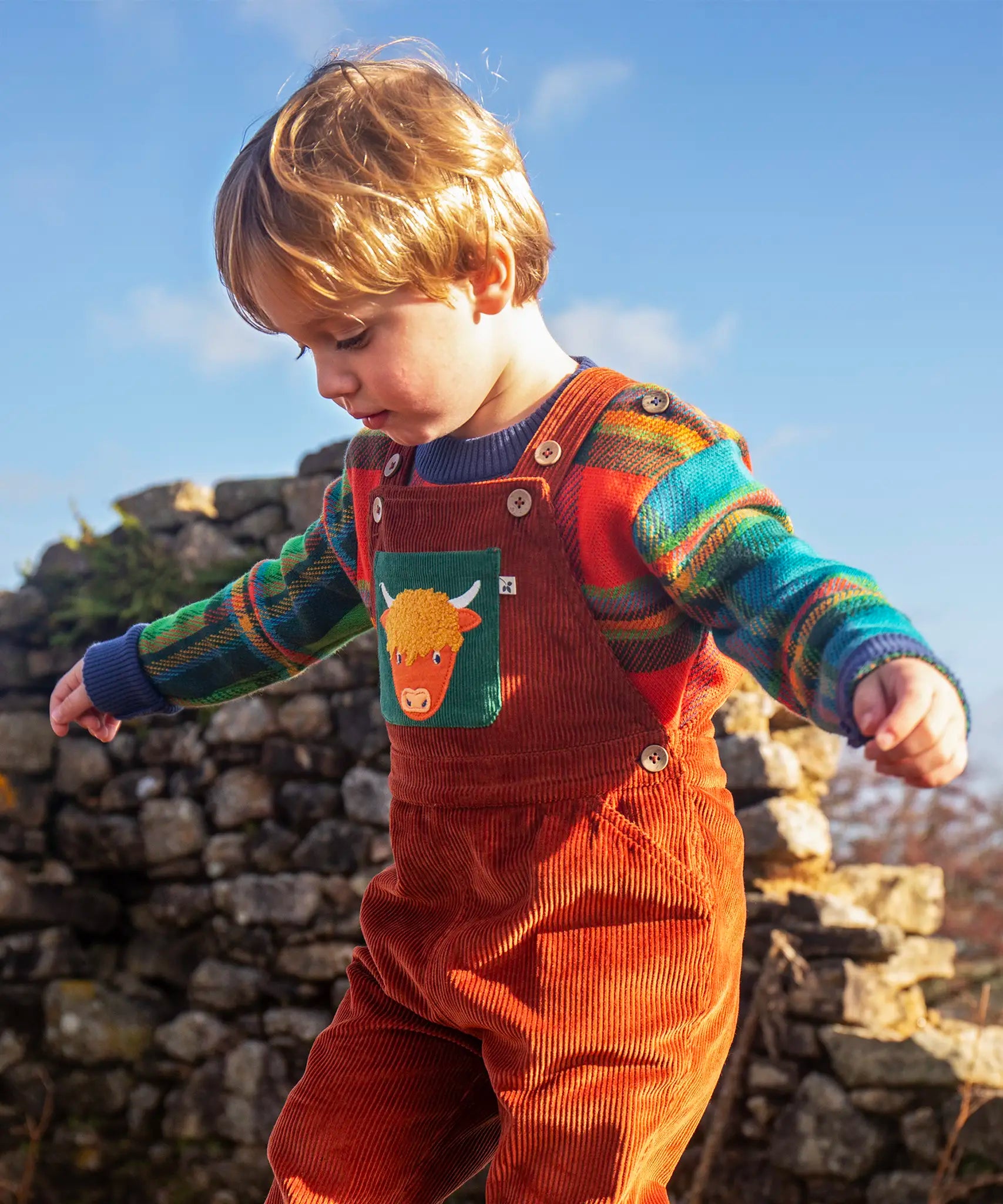 Child wearing the Frugi sebby brown rust organic cotton cord dungaree with a green chest pocket featuring a highland cow applique with a knitted jumper underneath.