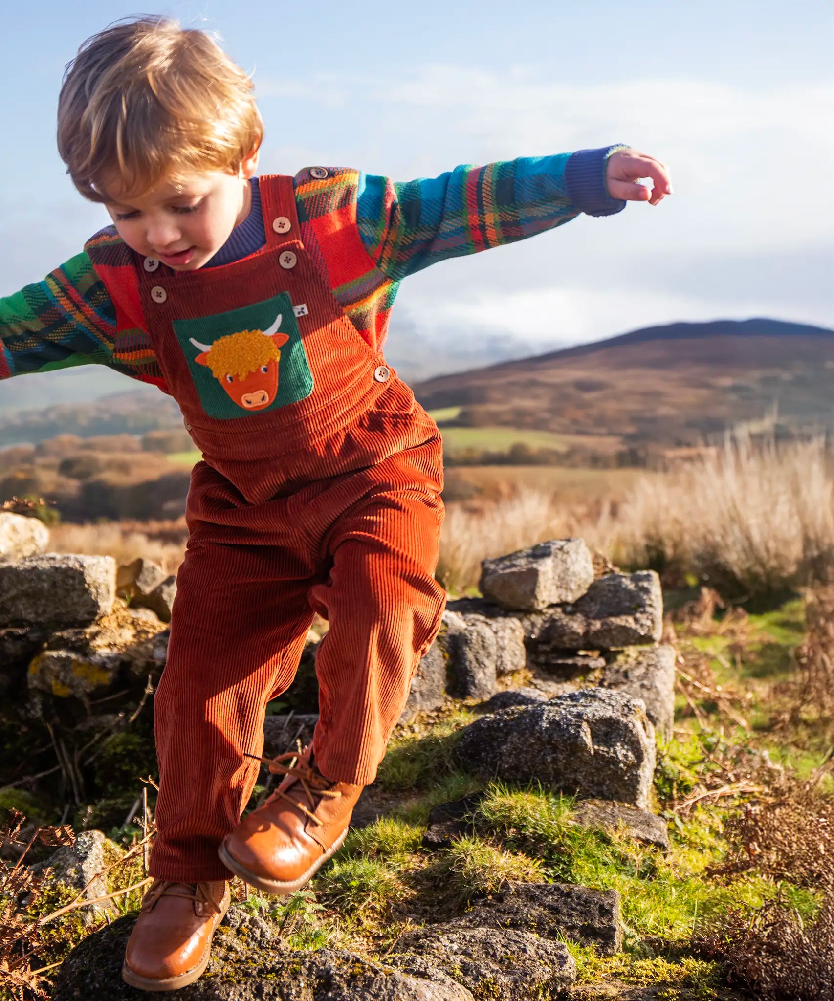 Child wearing the Frugi sebby brown rust organic cotton cord dungaree with a green chest pocket featuring a highland cow applique with a knitted jumper underneath.