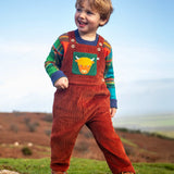 Child wearing the Frugi sebby brown rust organic cotton cord dungaree with a green chest pocket featuring a highland cow applique with a knitted jumper underneath.