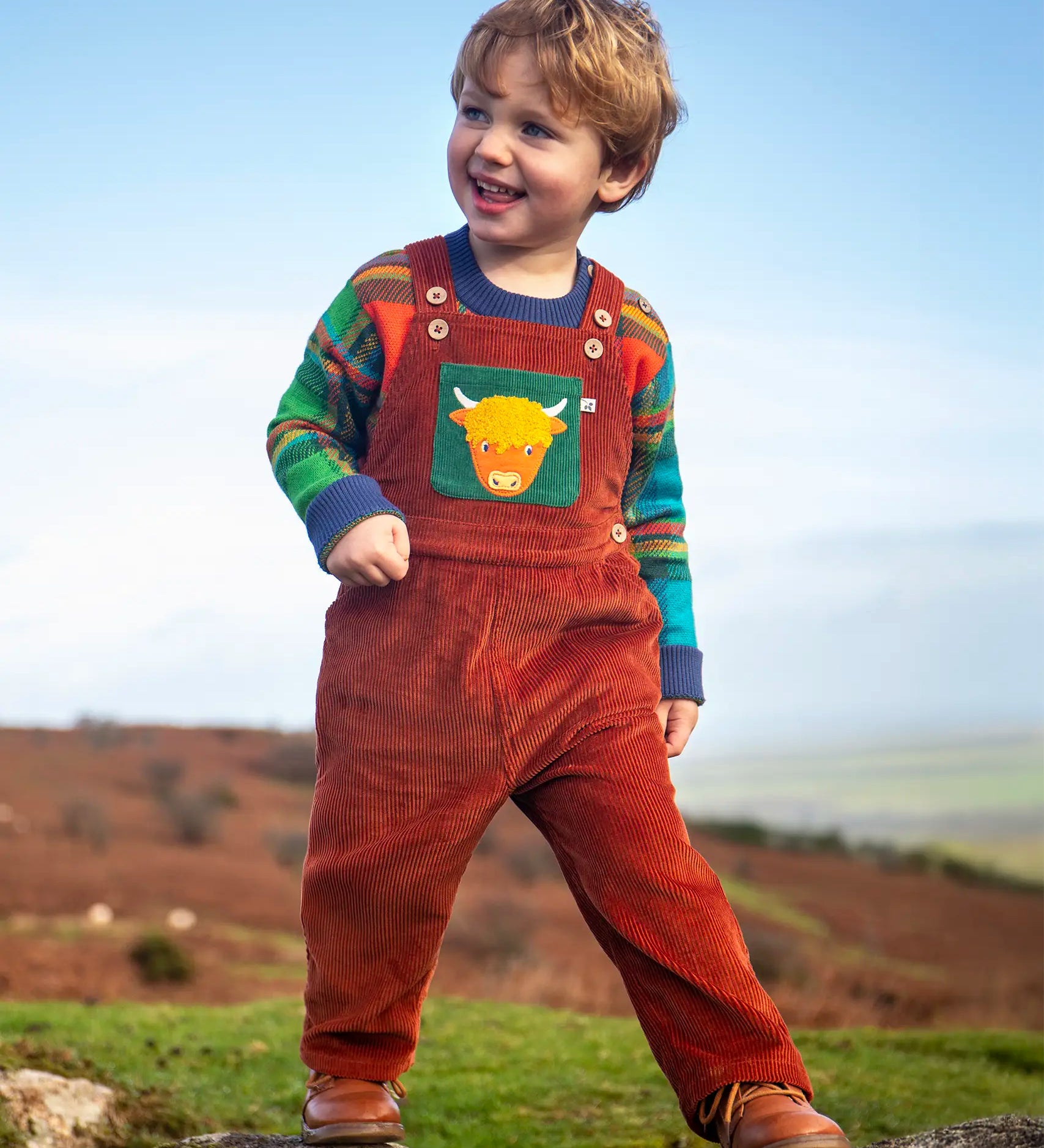 Child wearing the Frugi sebby brown rust organic cotton cord dungaree with a green chest pocket featuring a highland cow applique with a knitted jumper underneath.
