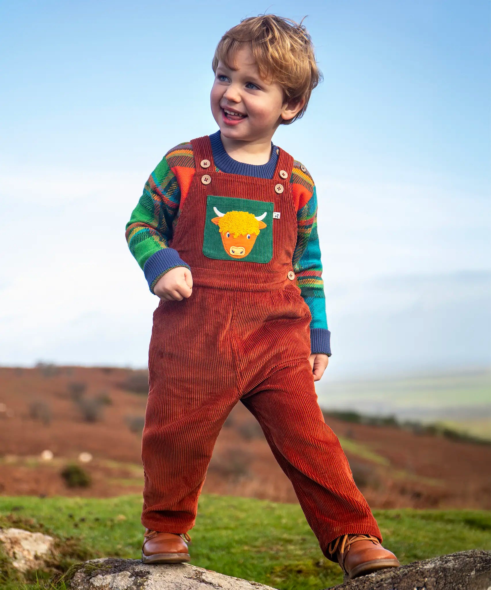 Child wearing the Frugi sebby brown rust organic cotton cord dungaree with a green chest pocket featuring a highland cow applique with a knitted jumper underneath.