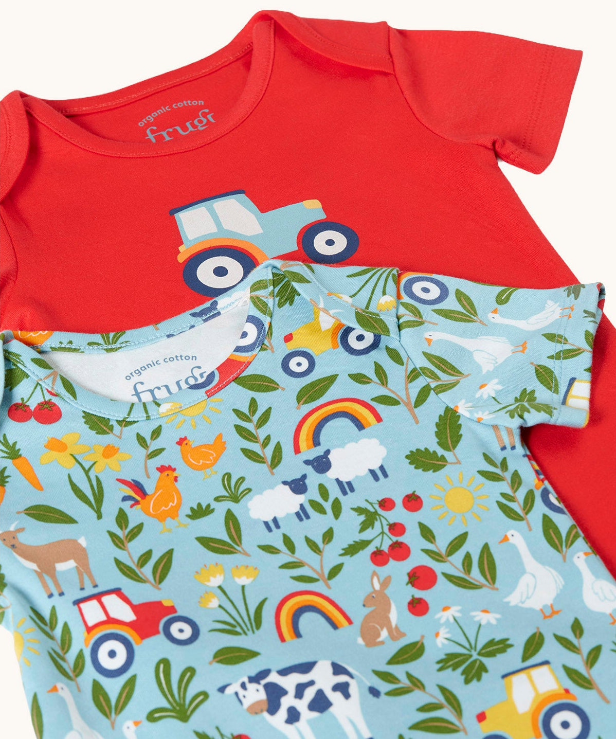 Close up of the front of the A two pack of Frugi short sleeve baby vests laid out on top of each other. One with a plain red background and a blue tractor printed on the front the other had a on the farm themed all over print featuring farm animals, tractors, leaves and plants on a light blue background.