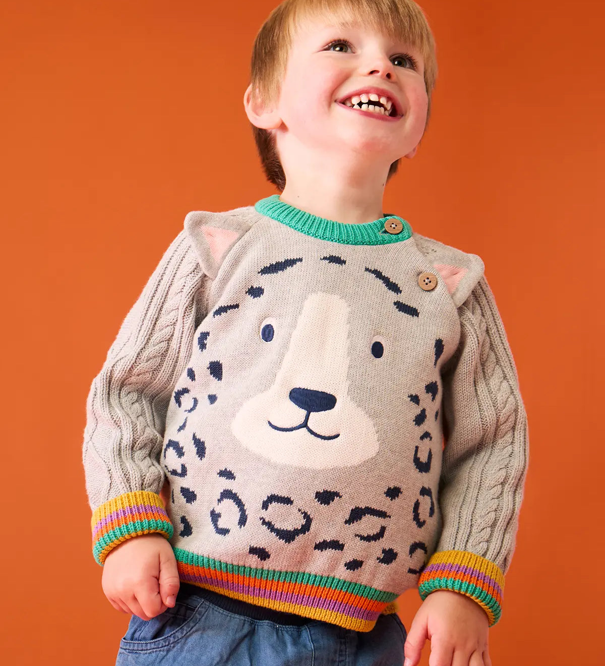 Child wearing the Frugi snow leopard organic cotton character knit jumper with 3d ears, green neck trim and rainbow arm cuffs and hemline. 