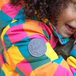 A close up of the Frugi kids snow and ski coat in rainbow check showing the grey brand circular patch stitched on the top of the arm on recycled polyester fabric