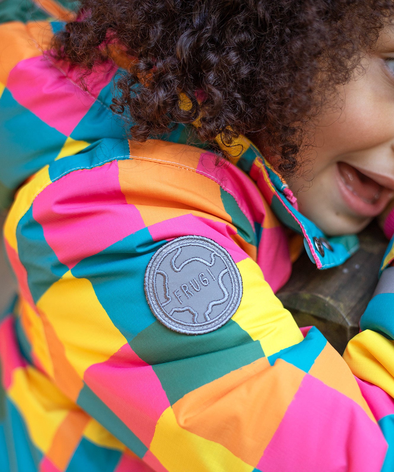 A close up of the Frugi kids snow and ski coat in rainbow check showing the grey brand circular patch stitched on the top of the arm on recycled polyester fabric