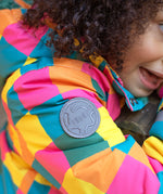 A close up of the Frugi kids snow and ski coat in rainbow check showing the grey brand circular patch stitched on the top of the arm on recycled polyester fabric