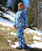A child wearing the Frugi Snow and Ski Salopettes in a Penguin Waddle design on a blue background with matching ski jacket. 