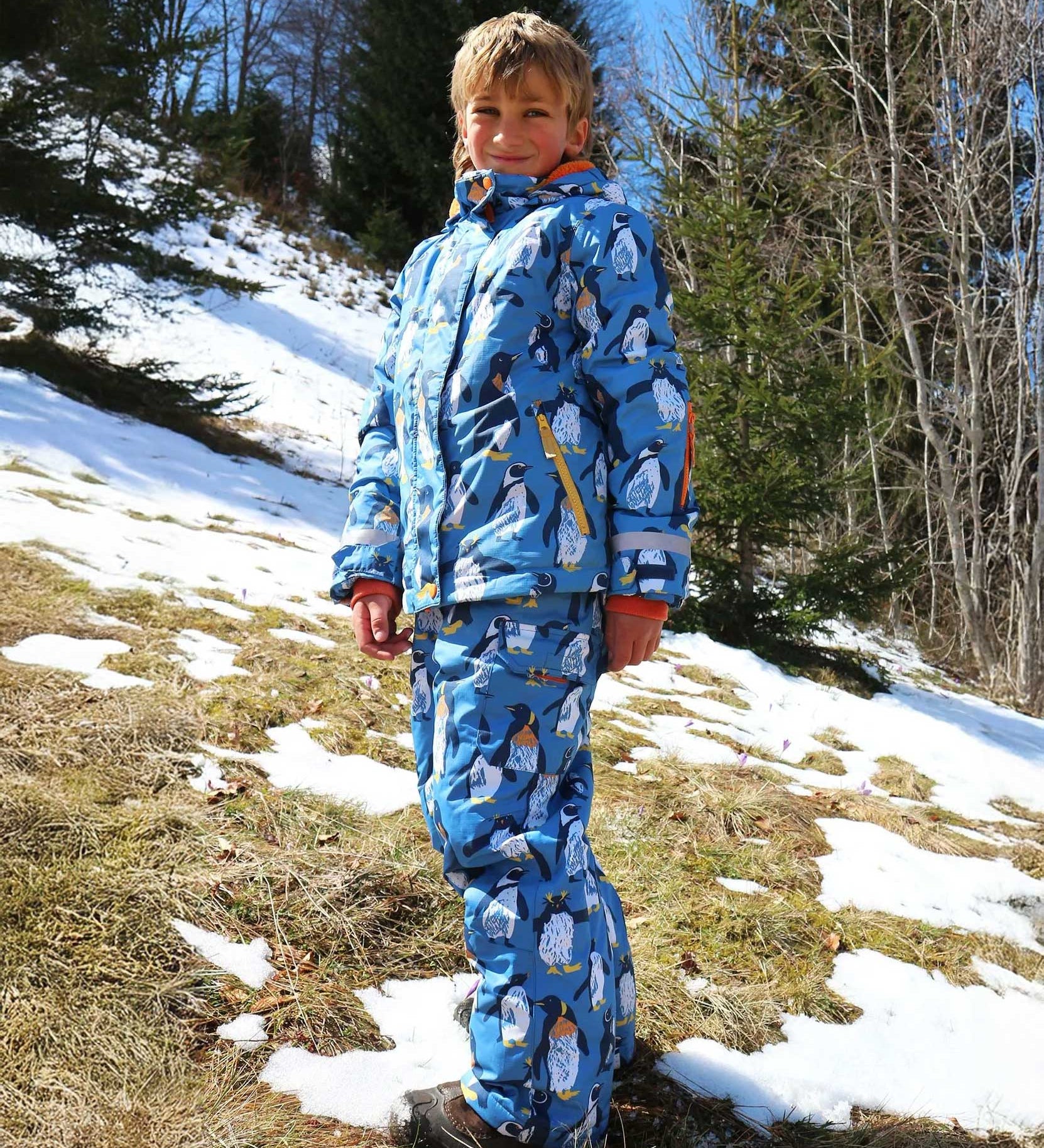 A child wearing the Frugi Snow and Ski Salopettes in a Penguin Waddle design on a blue background with matching ski jacket. 