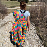 A child wearing the Frugi Snow and Ski Salopettes trousers in the Rainbow Check design and holding up the matching ski jacket. The design features squares in pink, orange, yellow, blue and green.