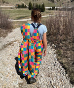 A child wearing the Frugi Snow and Ski Salopettes trousers in the Rainbow Check design and holding up the matching ski jacket. The design features squares in pink, orange, yellow, blue and green.