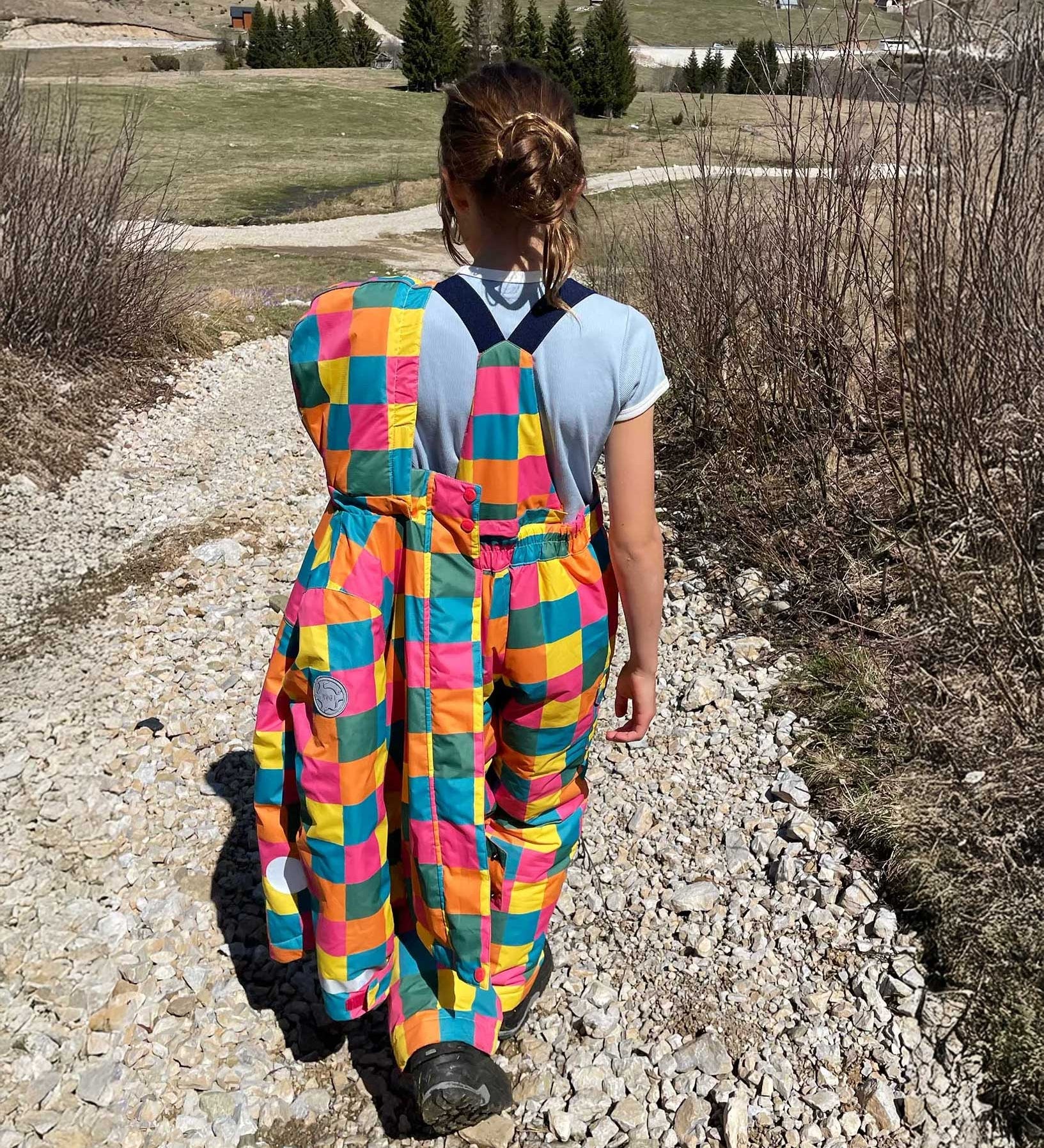 A child wearing the Frugi Snow and Ski Salopettes trousers in the Rainbow Check design and holding up the matching ski jacket. The design features squares in pink, orange, yellow, blue and green.