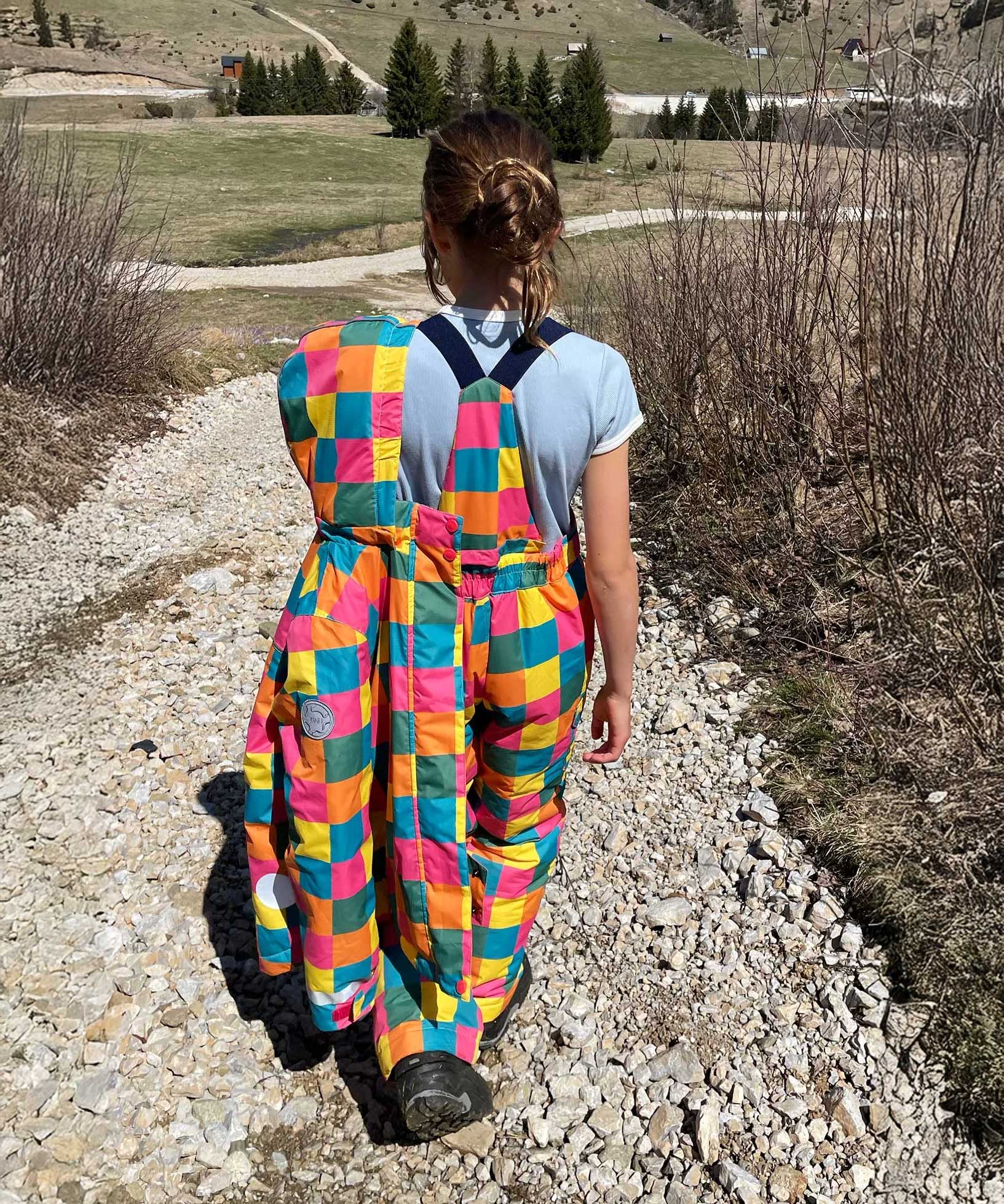 A child wearing the Frugi Snow and Ski Salopettes trousers in the Rainbow Check design and holding up the matching ski jacket. The design features squares in pink, orange, yellow, blue and green.