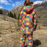 A child facing away from the camera wearing the Frugi Snow and Ski Salopettes in the Rainbow Check design with the matching ski jacket. The design features squares in pink, orange, yellow, blue and green.